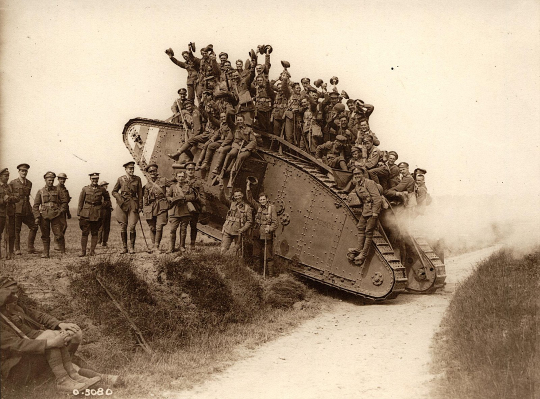 A large group of World War I soldiers, some waving hats, pose atop and beside a British tank on a dirt road; several other soldiers stand or sit nearby, with bushes in the foreground.