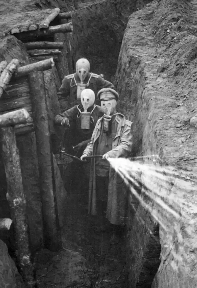 Three soldiers wearing early gas masks stand in a muddy World War I trench; one sprays a liquid, possibly disinfectant, while wooden reinforcements line the trench walls.