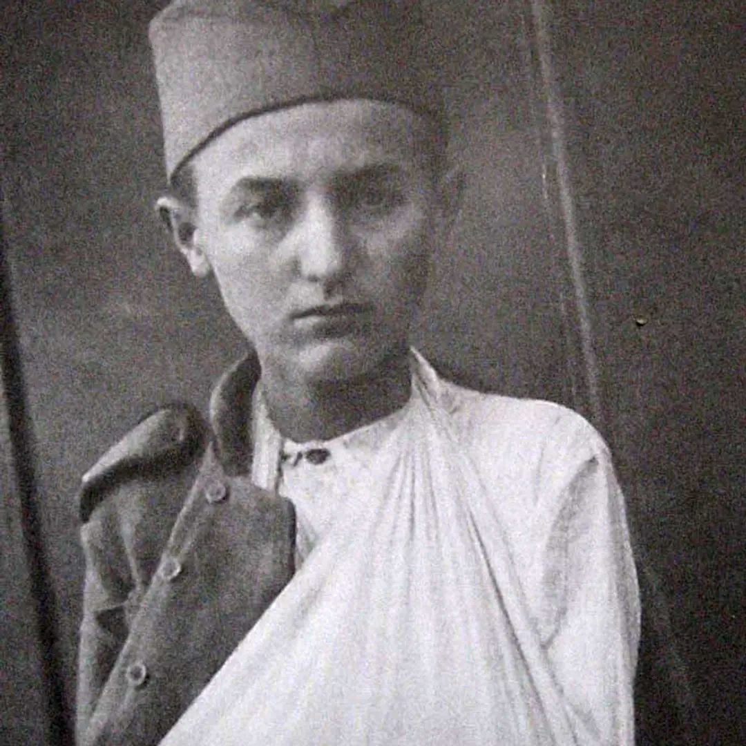 Black and white photo of a young boy wearing a uniform cap and jacket draped over one shoulder. His arm is in a sling, indicating an injury, and he looks directly at the camera with a serious expression.