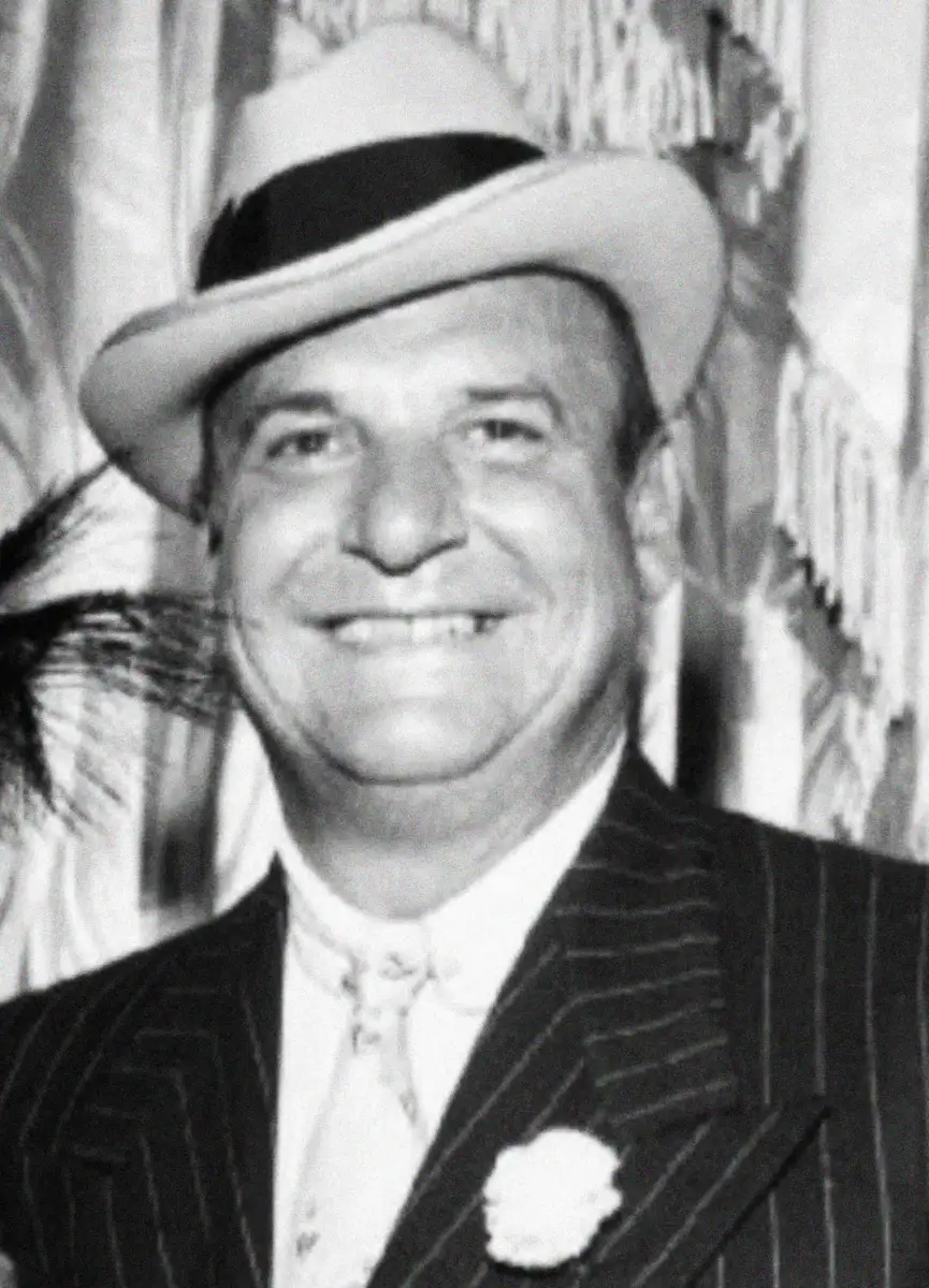 A smiling man in a pinstripe suit, patterned tie, and light-colored hat with a dark band, standing in front of fringed drapes.