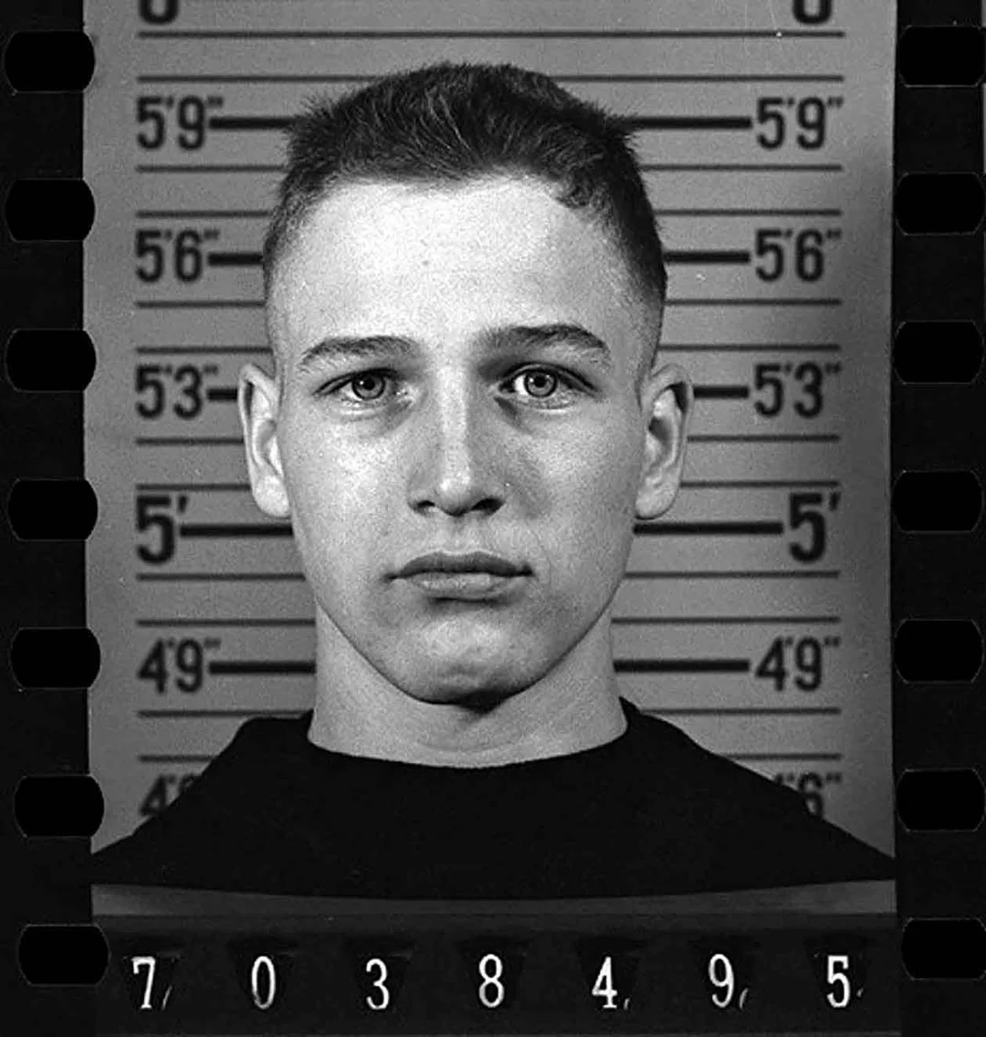 Black-and-white mugshot of a young man with short hair, standing in front of a height chart that measures up to 5'9". Numbers are visible at the bottom and sides of the image. The man is looking straight at the camera.