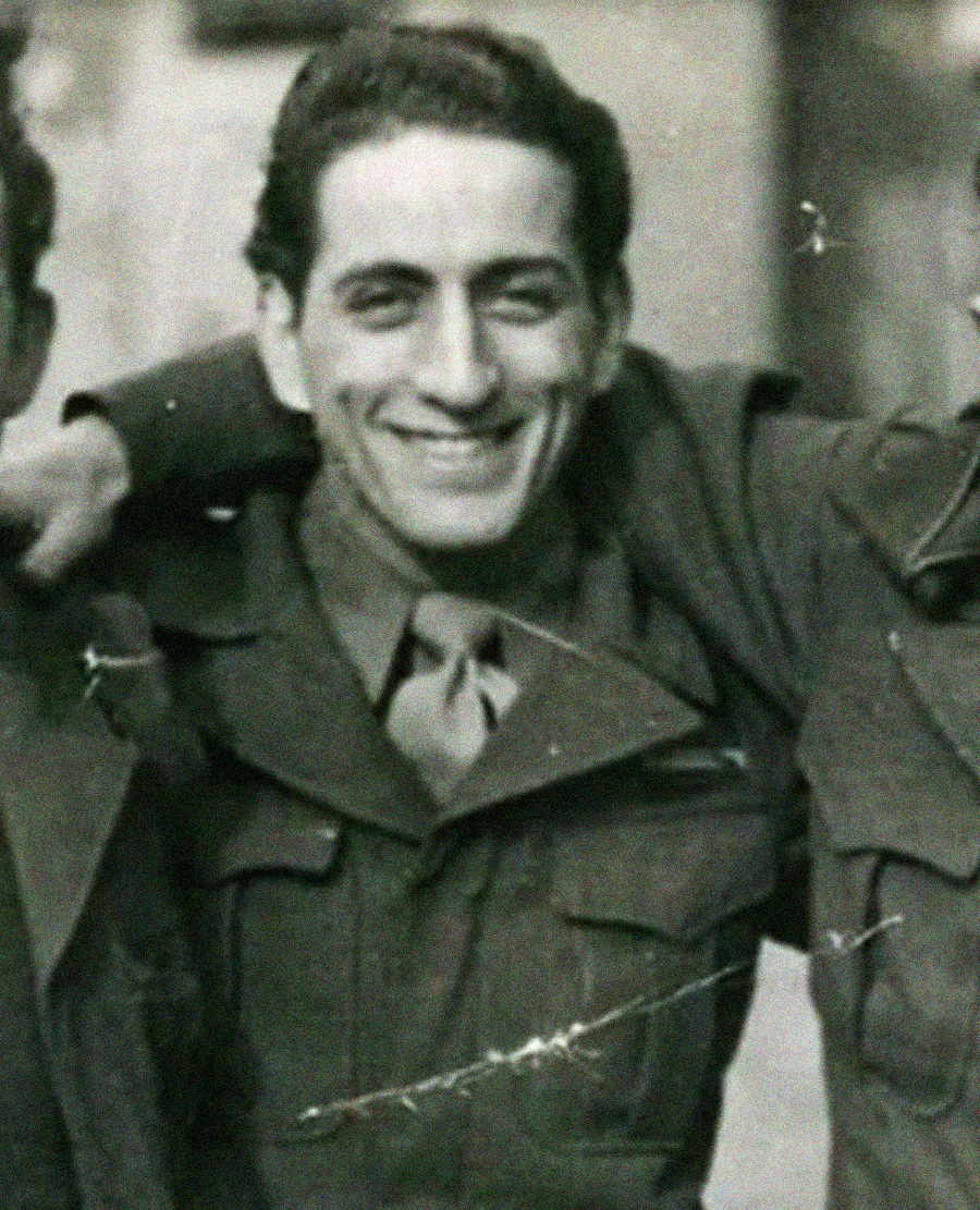 A young man in a military uniform smiles at the camera. The black-and-white photo is old and slightly scratched. The man has dark hair and is standing with his arms around others, who are partially visible.