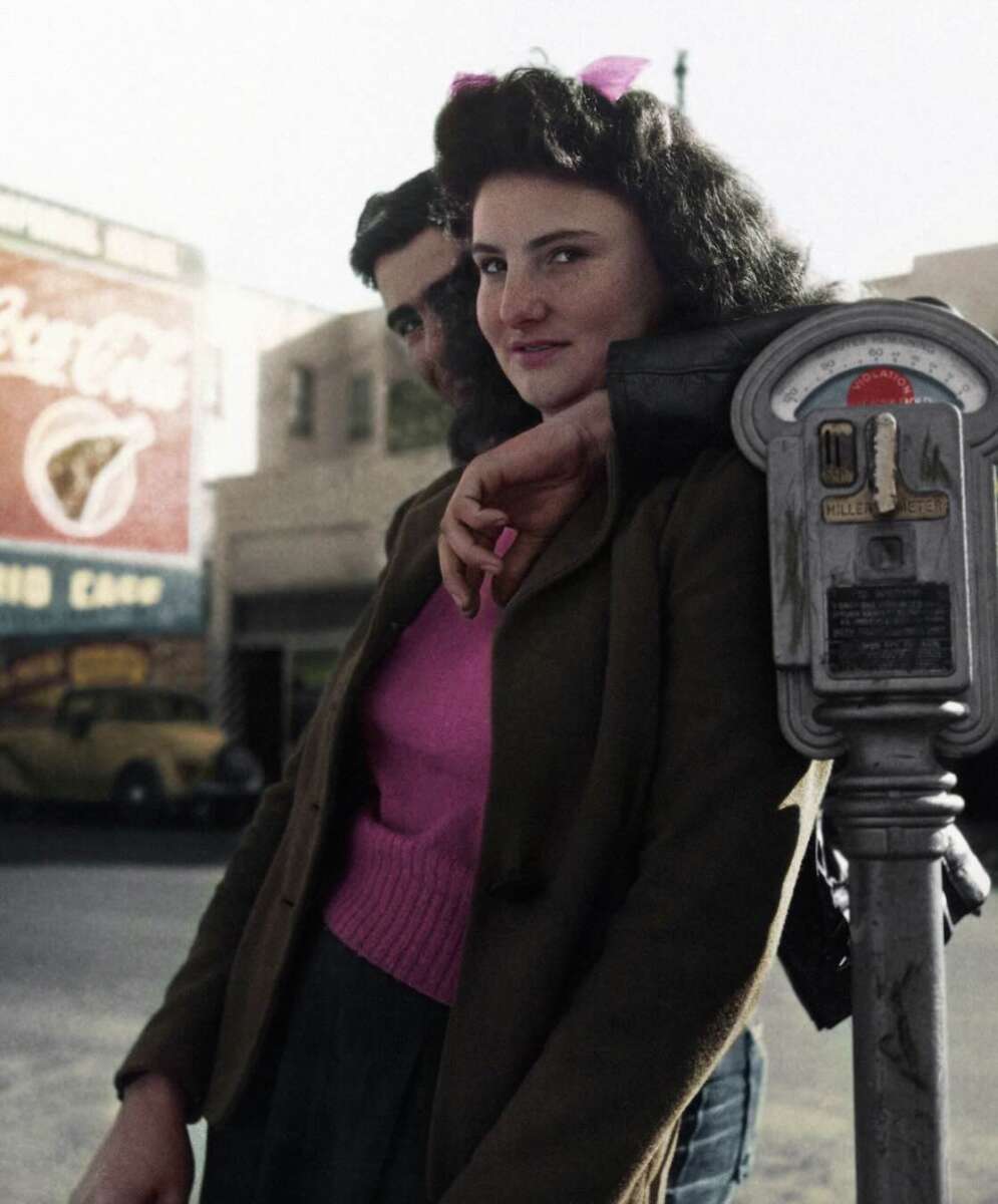 A woman in a pink sweater and dark jacket stands next to a man, leaning on a parking meter. She smiles slightly at the camera on a city street with old cars and a Coca-Cola billboard in the background.