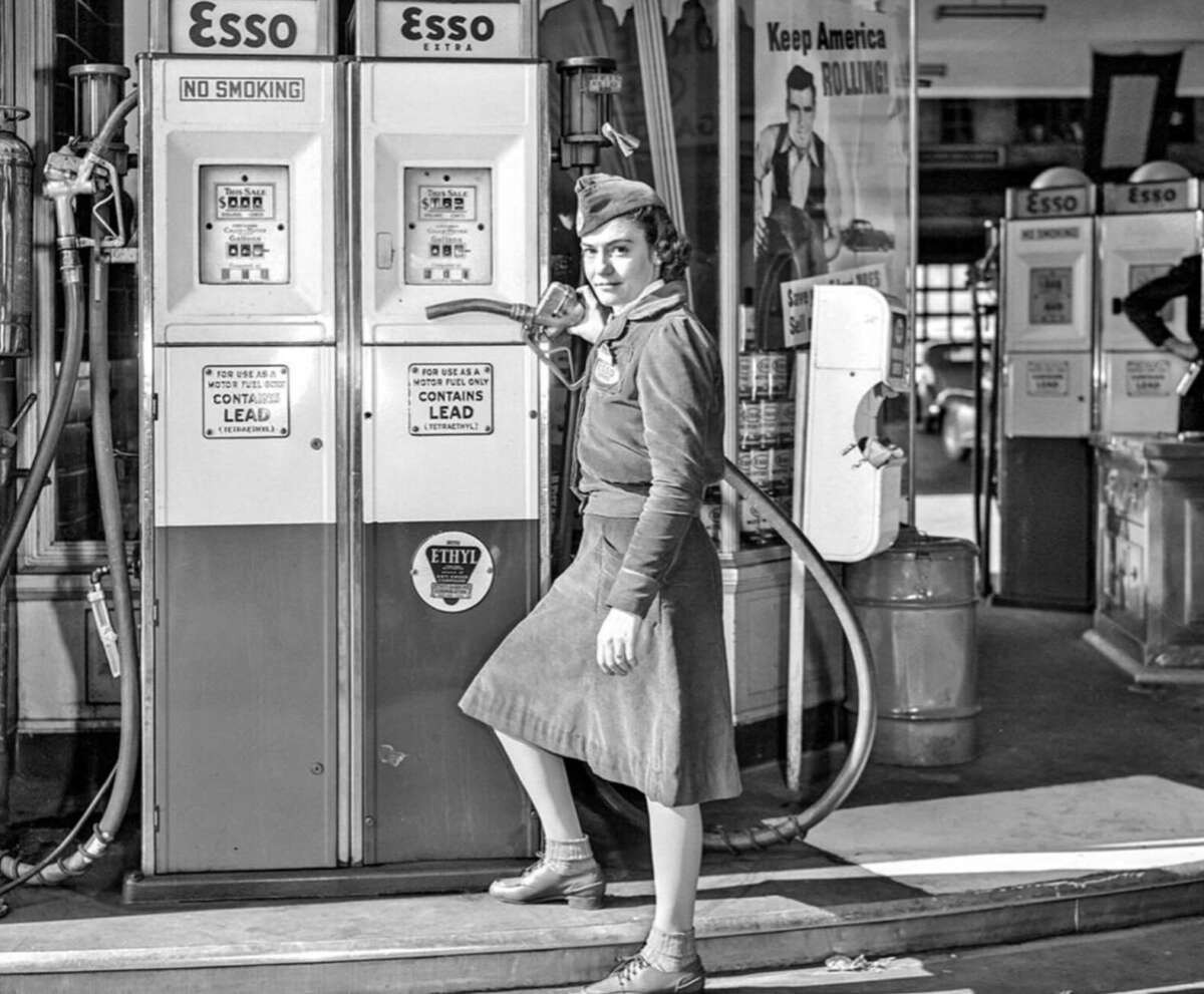 A woman in vintage clothing stands at an Esso gas pump, holding the nozzle. The setting appears to be an old-fashioned gas station, with signs reading “No Smoking” and “Contains Lead.”