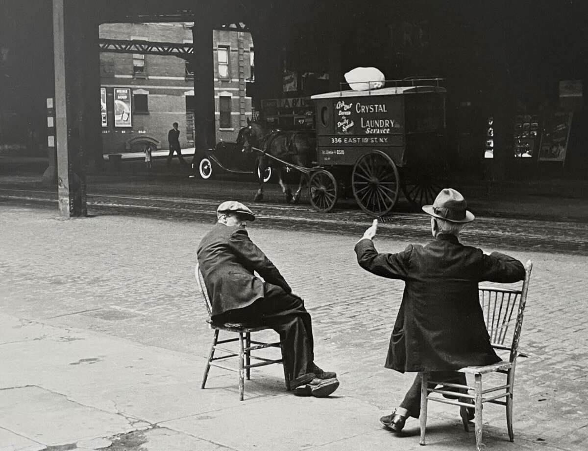Two men sit on wooden chairs on a sidewalk, facing a street. One gestures with his hand while the other looks on. An old-fashioned horse-drawn laundry wagon is parked across the street under a bridge.