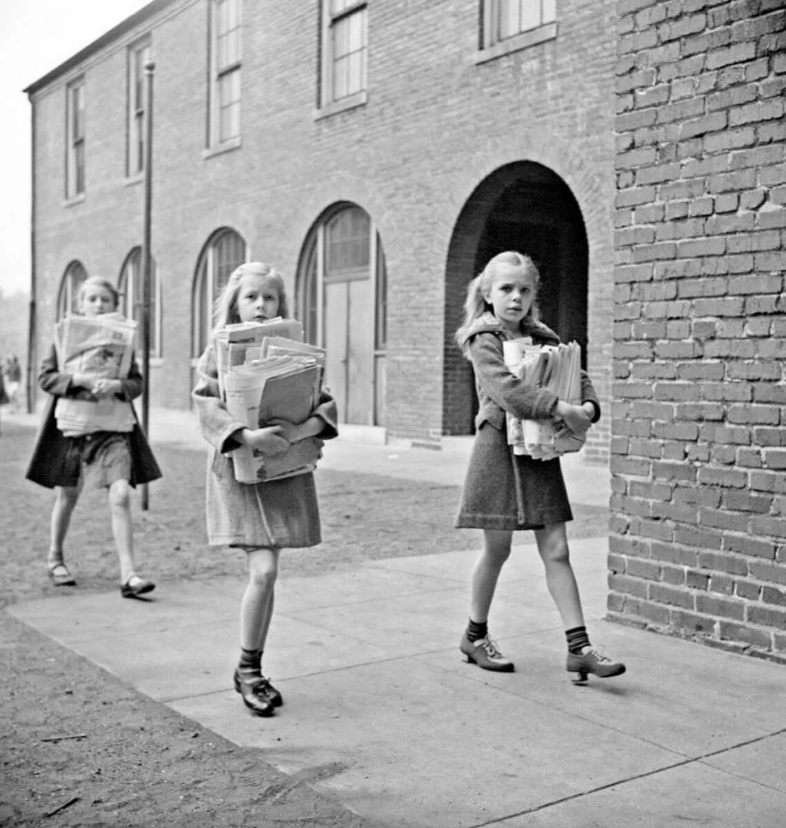 Three young girls walk on a sidewalk beside a brick building, each holding a large stack of newspapers in their arms. They wear coats and skirts, and have long hair, appearing focused on their tasks.