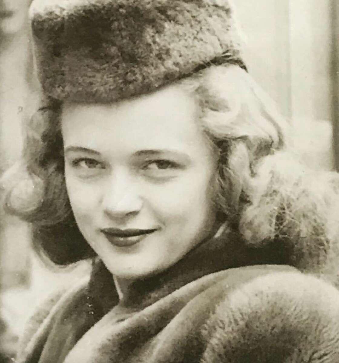 A young woman with wavy hair smiles softly, wearing a fur hat and coat in a black-and-white vintage photograph.