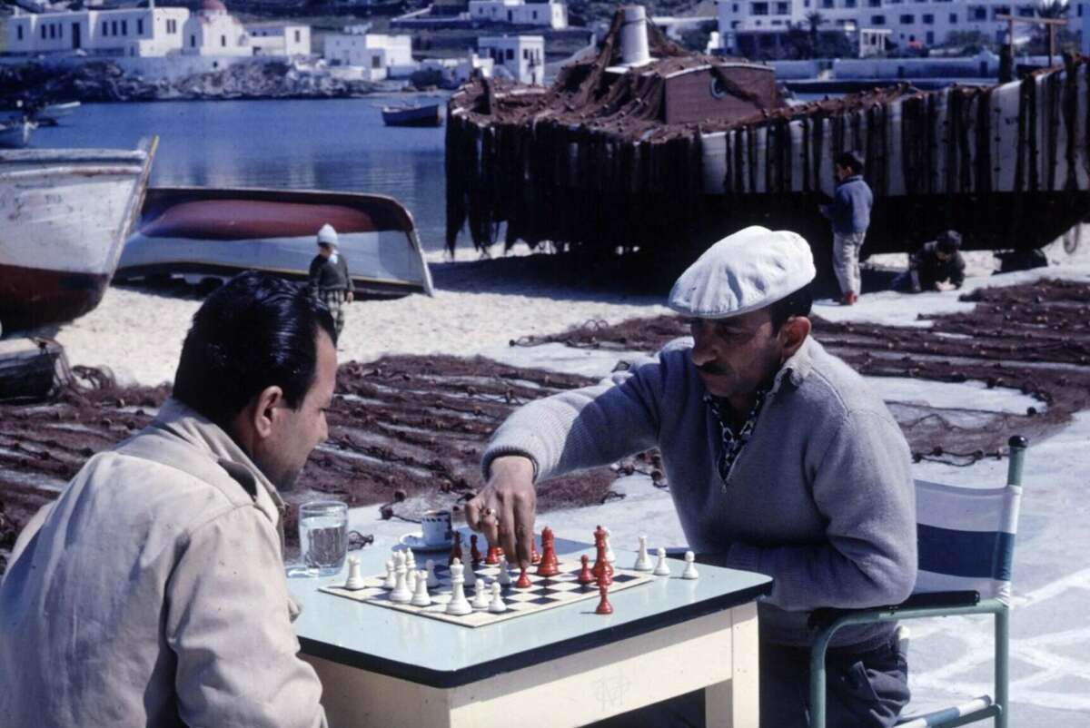 Two men play chess at a table on a sandy waterfront, with boats, fishing nets, and seaside buildings in the background. One man is making a move while the other watches.