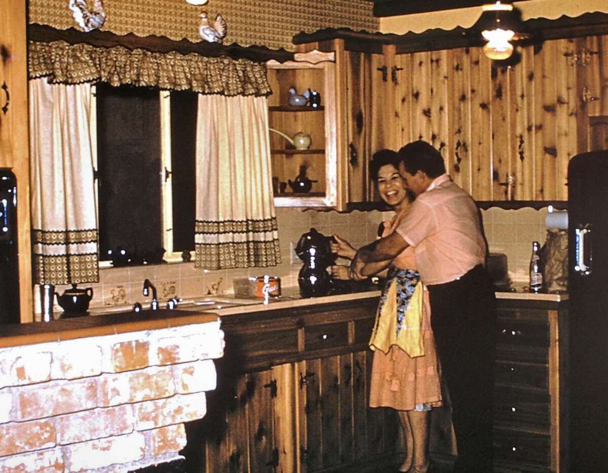 A woman in a dress and apron smiles as a man hugs her in a retro-style wooden kitchen with patterned wallpaper, a brick counter, and vintage appliances.
