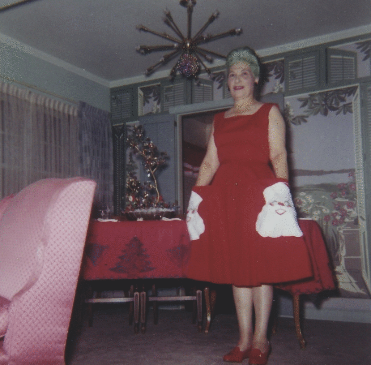 A woman in a festive red dress with Santa face pockets stands smiling in a decorated room with holiday decor, including a table covered with a red Christmas tablecloth and a silver Christmas tree.