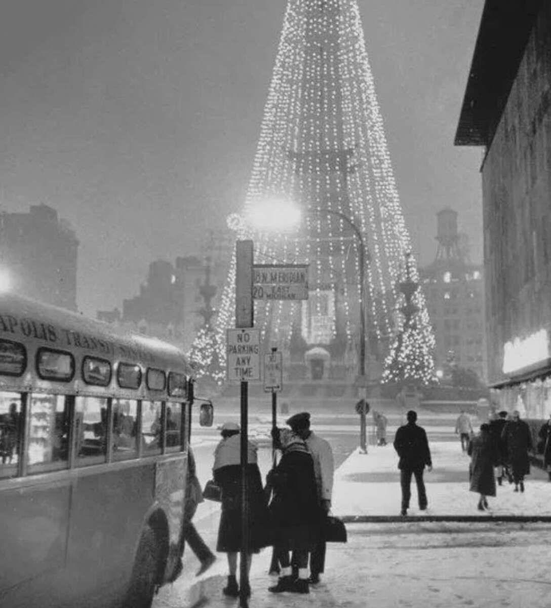 Black and white photo of people standing near a bus stop on a snowy street at night, with a large, illuminated Christmas tree in the background and city buildings surrounding the scene.