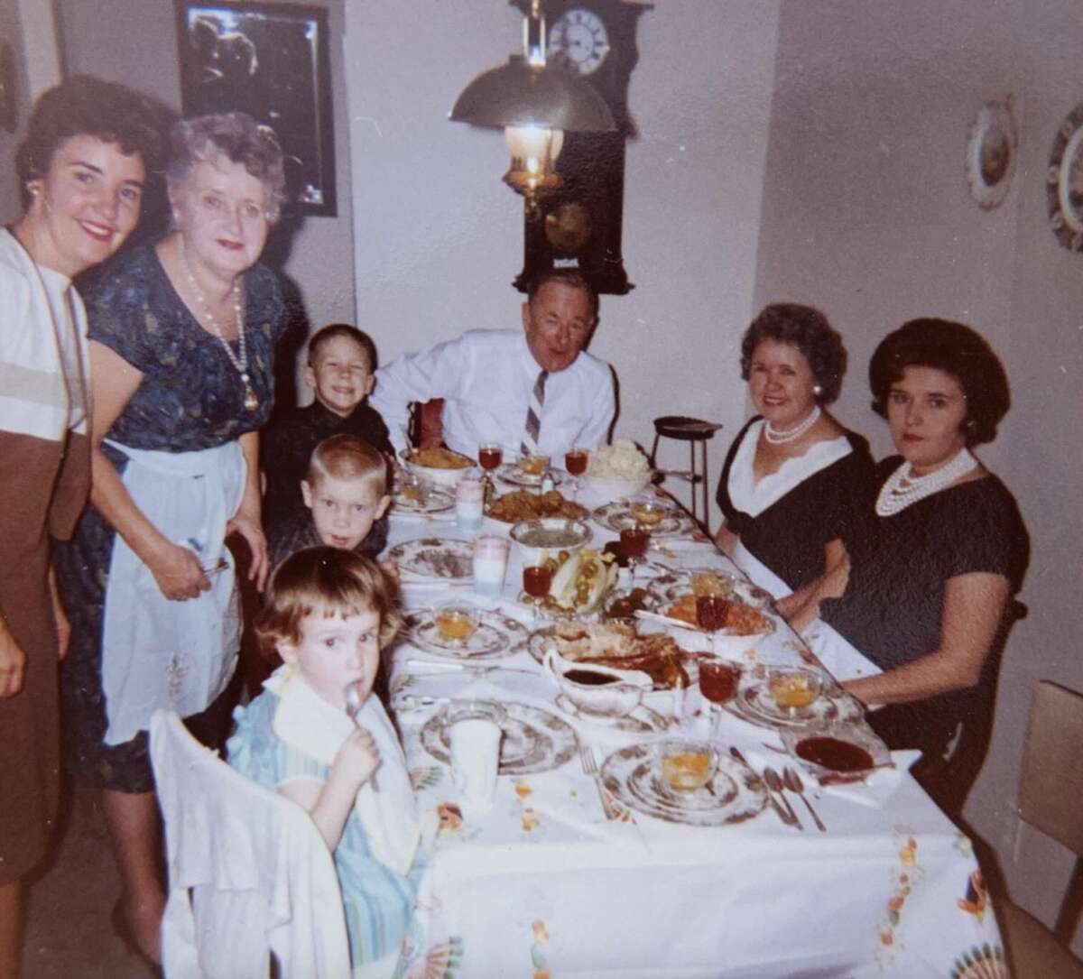 A vintage photo of seven people, both adults and children, gathered around a dining table set with plates of food, drinks, and desserts. The group is smiling, and the room is warmly lit with a clock on the wall behind them.