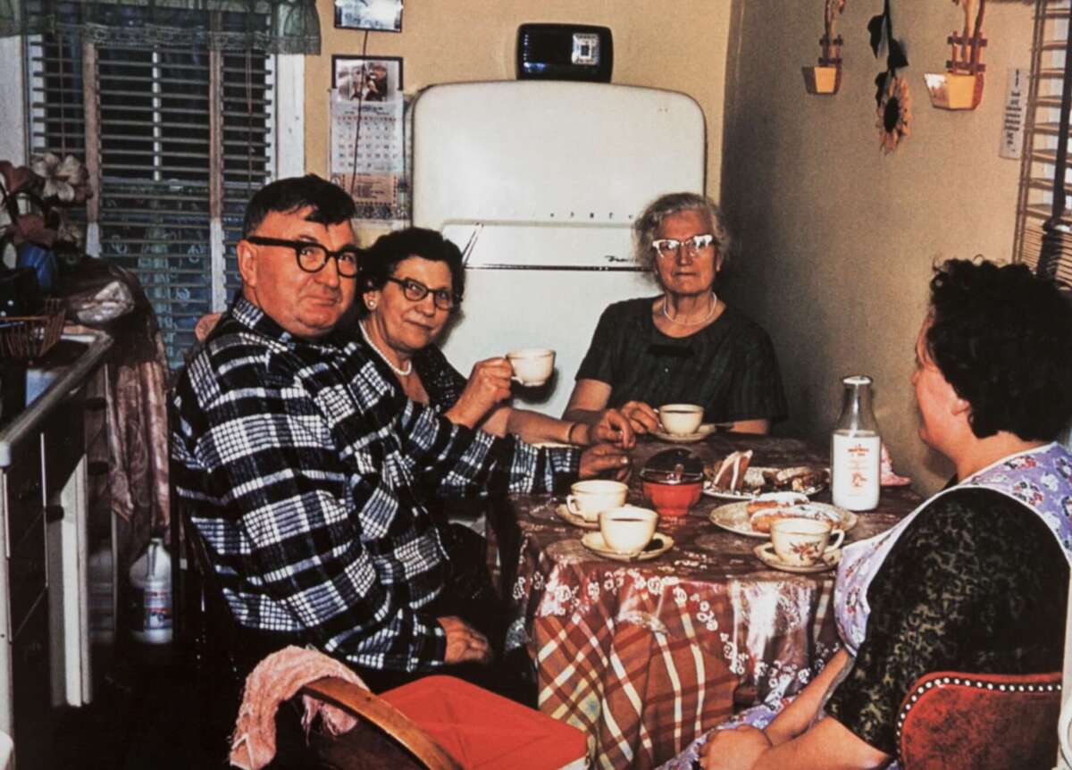 Four older adults sit around a kitchen table with tea cups, pastries, and a bottle of milk. The kitchen has a vintage refrigerator and patterned curtains. The mood appears relaxed and social.