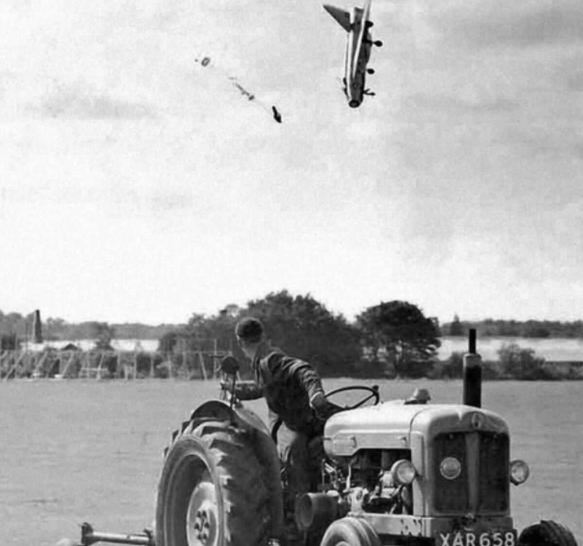 A black-and-white photo shows a man on a tractor looking up as a military plane crashes nose-first in the sky above, trailing smoke and debris over a rural landscape.