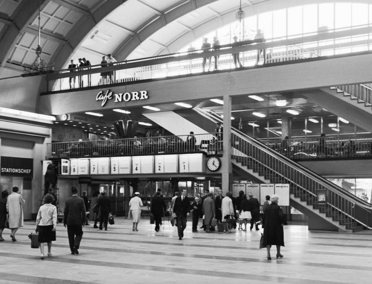Busy indoor train station in black and white with people walking, a staircase, large windows, a clock, and a sign for Café Norr. Some people stand, others walk, and several are visible on an upper balcony.