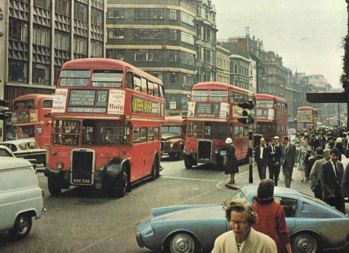 A busy city street scene with several red double-decker buses, crowded sidewalks with pedestrians, and various cars, including a blue sports car in the foreground. The buildings are tall and urban in style.