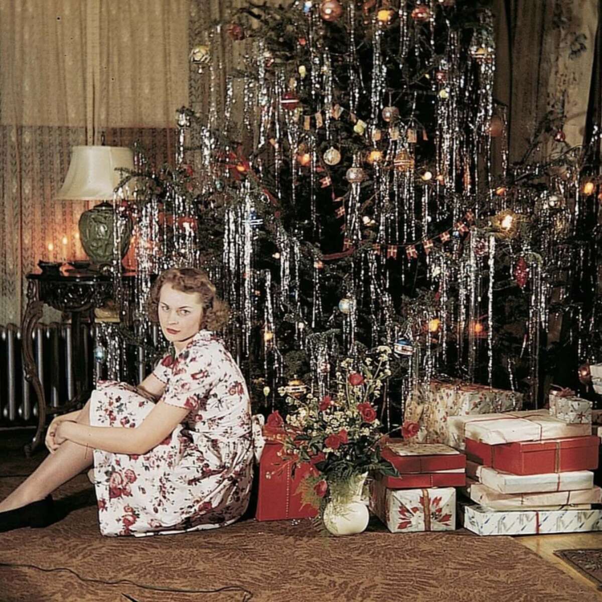 A woman in a floral dress sits on the floor beside a decorated Christmas tree with tinsel and ornaments. Wrapped presents and a bouquet of flowers are arranged around the base of the tree in a warmly lit room.