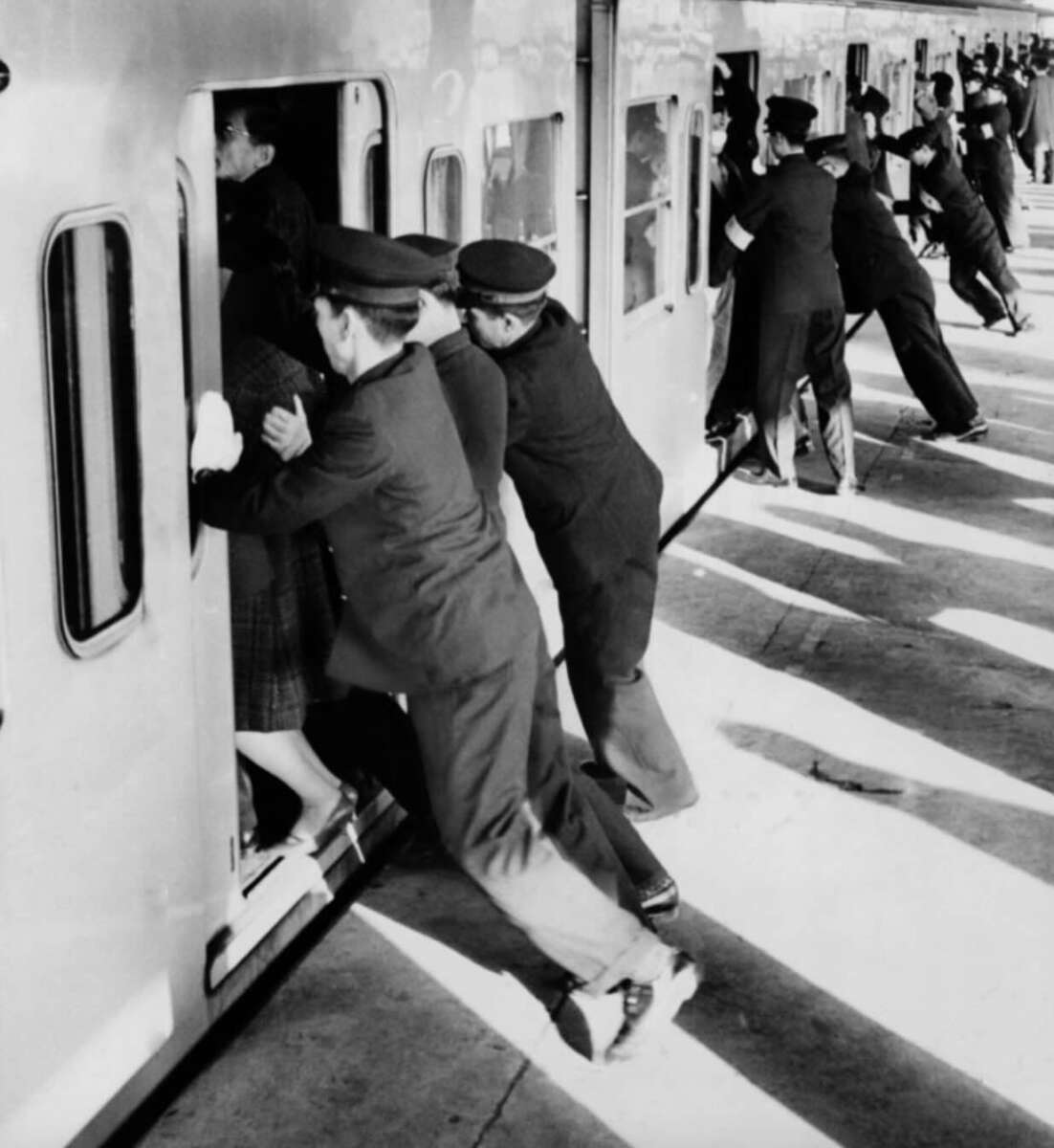 Train station staff in uniforms push passengers into a crowded train car, helping them fit inside before the doors close. The platform is filled with several staff members assisting along the length of the train.