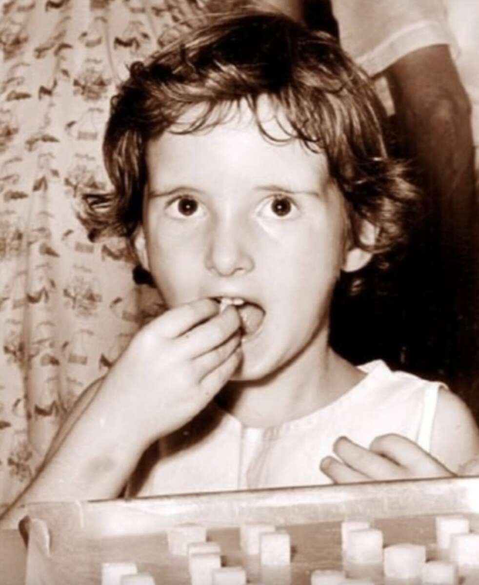 A young child with short curly hair eats sugar cubes from a tray, looking directly at the camera with wide eyes. The image is in black and white, and people are blurred in the background.