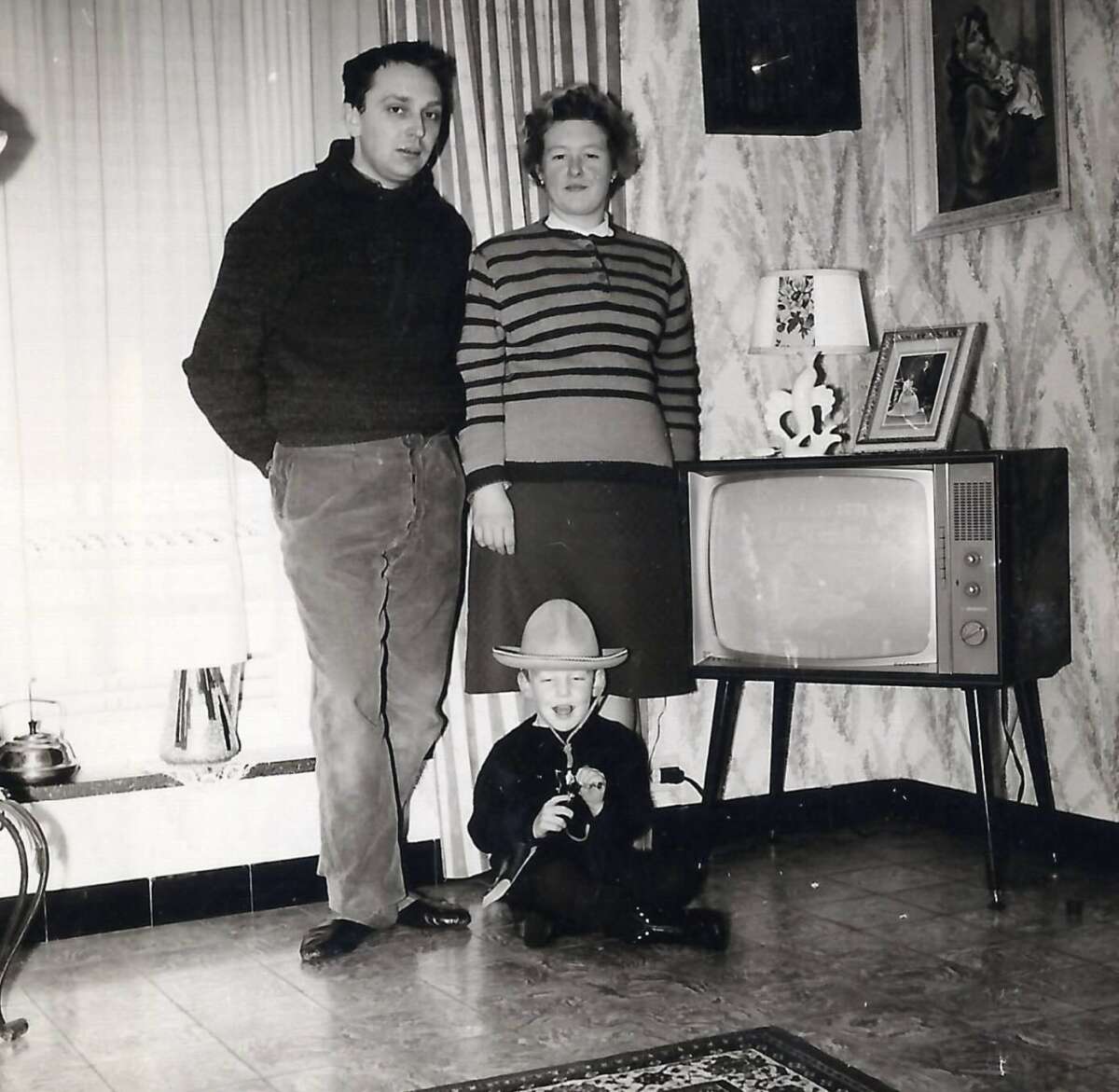 A man and woman stand beside an old-fashioned TV in a vintage living room, while a young child in a cowboy hat sits on the floor, smiling and holding a toy. The room has patterned wallpaper and framed photos.