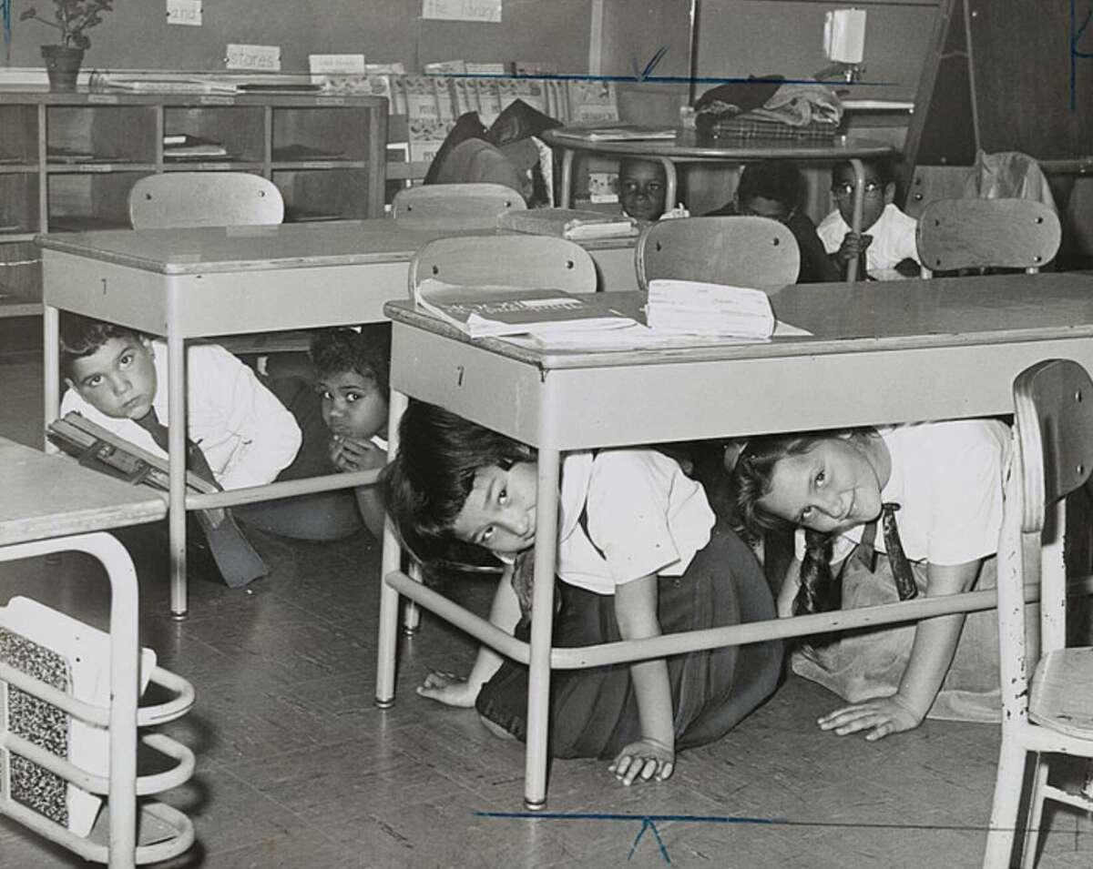 Children crouch under their desks in a classroom during a drill. Books and supplies are on the desks, and other students are visible seated in the background. The scene appears to be from a mid-20th century school.