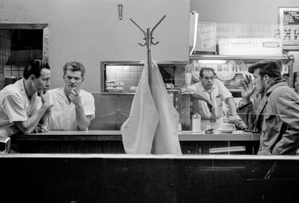 Black-and-white photo of three men behind a diner counter and one man sitting on a stool, all engaged in conversation. A coat rack and coat partially obscure the scene. The setting appears casual and retro.