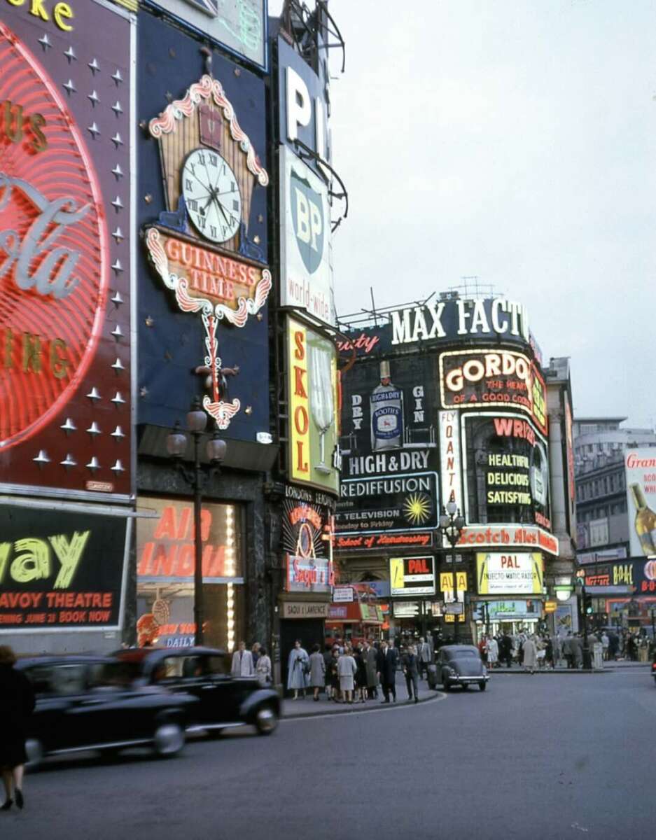 A busy city street scene from the mid-20th century features colorful neon billboards and advertisements above pedestrians and vintage cars, including signs for Guinness, BP, Coca-Cola, and Gordon’s Gin.