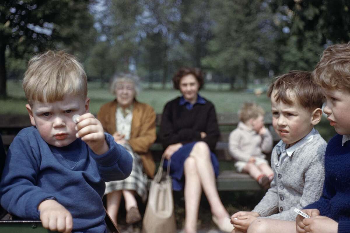 Three young boys sit on a park bench in the foreground, with two women and a small child blurred in the background. Trees and greenery fill the background. The mood appears casual and relaxed.