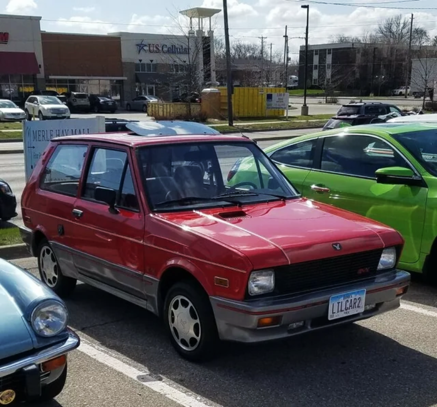 A small red vintage hatchback car with a “LIL CAR2” license plate is parked in a lot between other cars on a sunny day. Buildings and a US Cellular store are visible in the background.