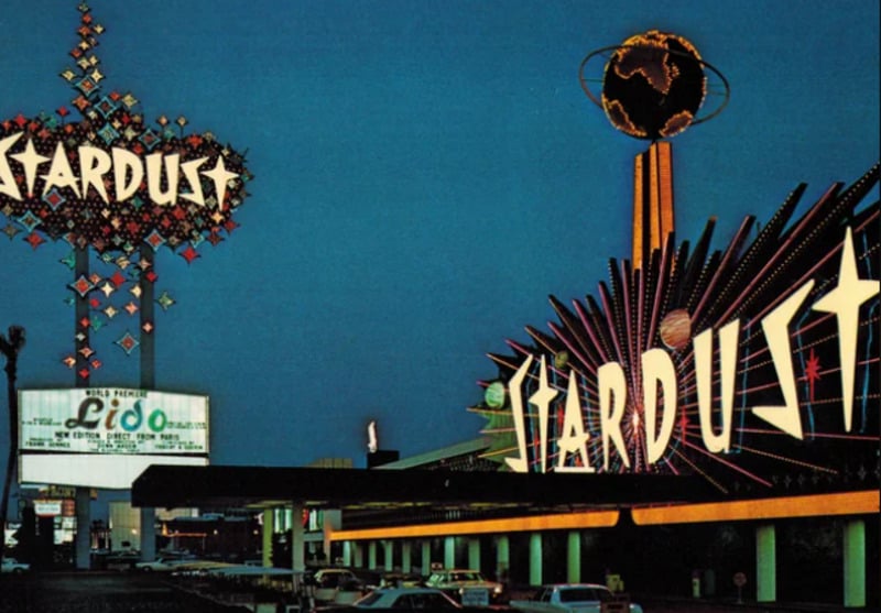 Neon signs for the Stardust hotel and casino glow brightly at night in Las Vegas, with bold, colorful lights and a globe atop one sign. Several cars are parked in front, and a Lido show billboard is visible.
