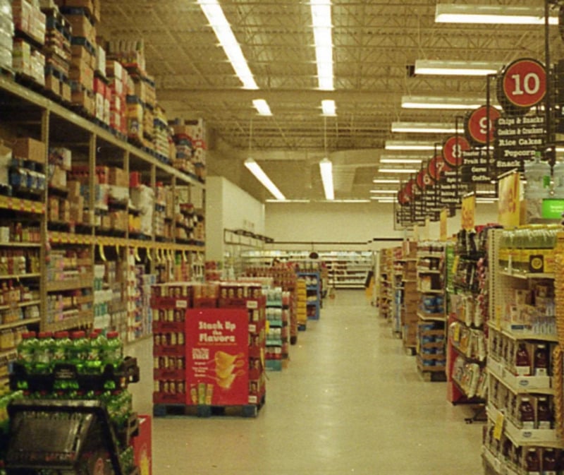 A grocery store aisle with shelves stocked with food and drinks, overhead fluorescent lighting, and red numbered aisle signs hanging from the ceiling.