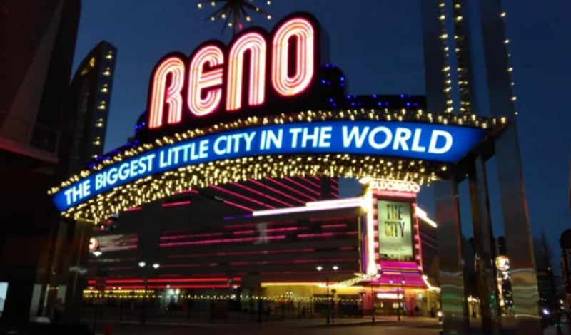 The illuminated Reno arch at night displays the slogan "The Biggest Little City in the World" in bright lights, with a casino and additional neon signs glowing in the background.