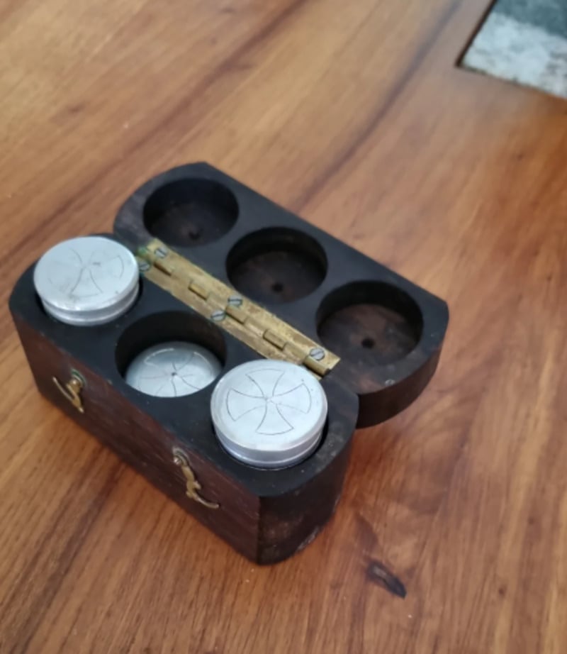 A small wooden box with a brass hinge, holding four round metal containers, sits open on a wooden surface. The containers have simple geometric designs engraved on their lids.