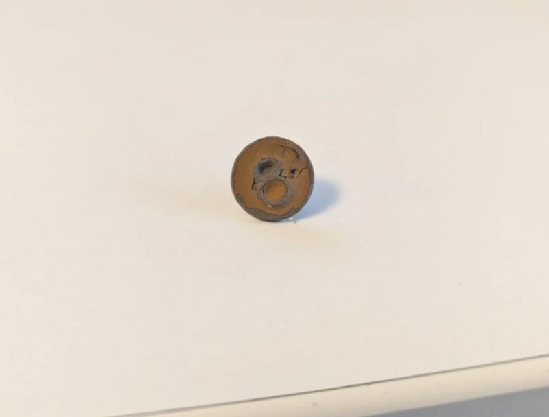 A small, round, brownish object with two holes sits on a white surface. The object appears aged and cracked, possibly made of metal or ceramic.