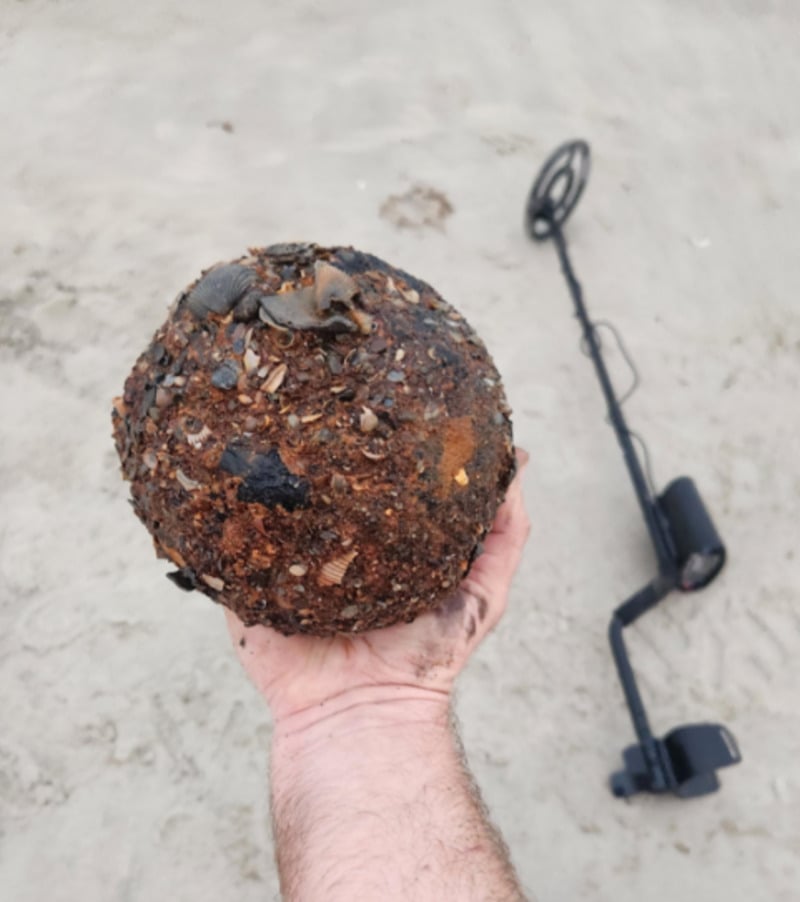 A hand holds a large, round, rusted object covered in sand and shells on a beach; a metal detector rests on the sand in the background.