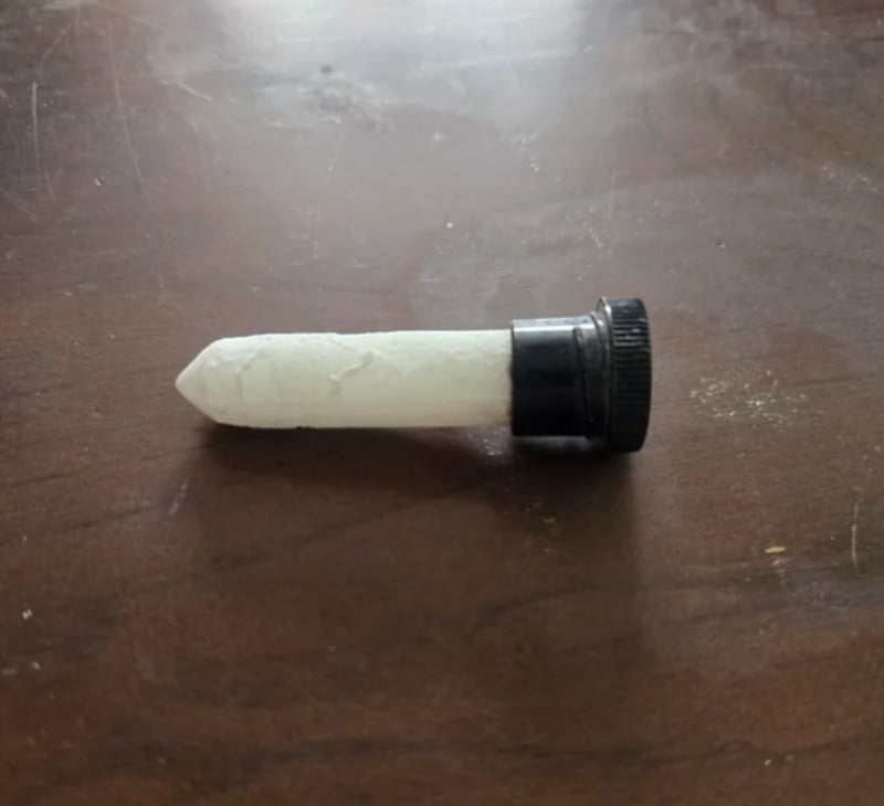 A white chalk stick with one pointed end is inserted into a black plastic bottle cap, lying on a dark wooden surface.