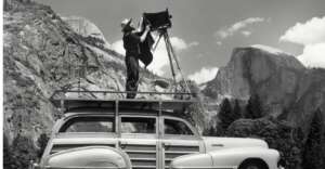A photographer stands on the roof of a vintage car, adjusting a large camera on a tripod, with mountains and trees in the background under a partly cloudy sky.