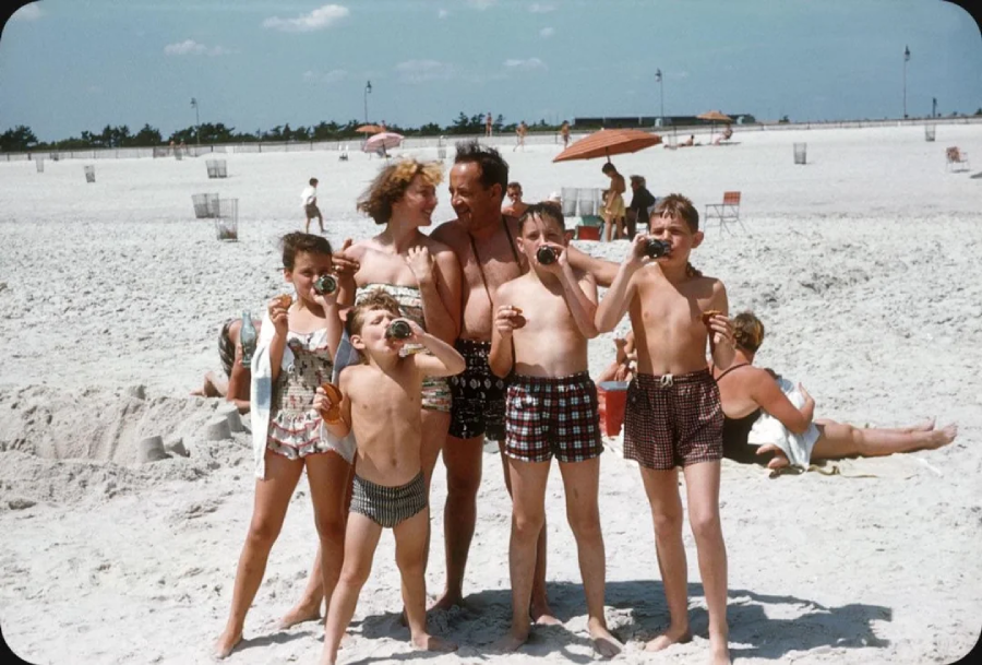 A group of five children and one adult, all in swimsuits, stand together smiling and eating popsicles on a sandy beach with scattered umbrellas, chairs, and people in the background under a blue sky.