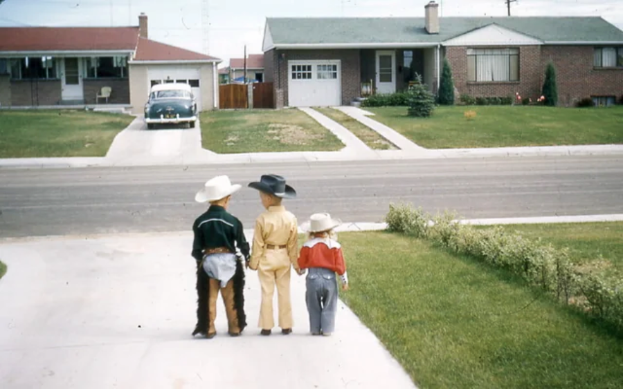 Three young children dressed in cowboy outfits stand on a suburban driveway, facing a quiet street. Across the road are two houses with garages and a vintage car parked in front of one.