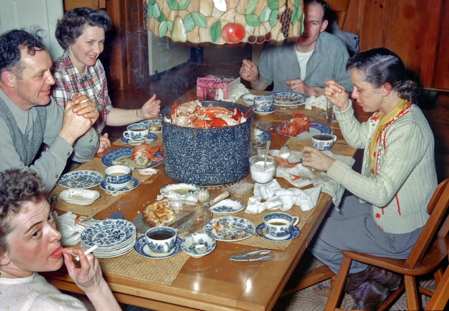 Five people sit around a dining table, sharing a meal with a large pot of cooked crabs at the center. The table is set with blue and white dishes, cups, and various foods. Steam rises from the pot as they eat together.