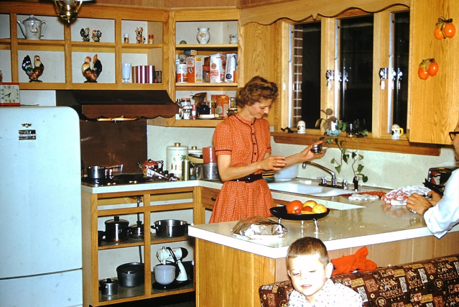 A woman in a red polka-dot dress stands at a kitchen sink, washing dishes. A child sits on a couch in the foreground. The kitchen has wooden cabinets, open shelves with dishes and rooster figurines, and a bowl of fruit on the counter.