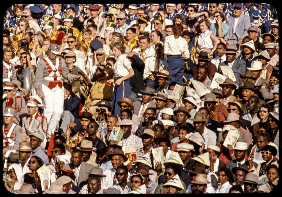 A large, diverse crowd sits closely packed in stadium bleachers, with some people wearing hats and formal clothing, and others in band uniforms. Many are watching attentively, holding programs, or interacting with one another.