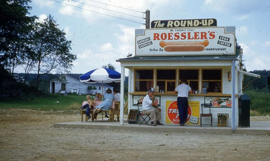 A vintage roadside hot dog stand called "The Round-Up" with a Roessler’s sign. Several people sit at tables under umbrellas, while one person stands at the counter. A rural landscape and small white building are visible in the background.