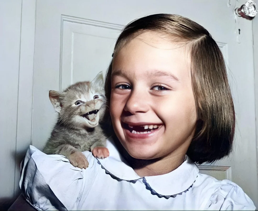A young girl with short brown hair and a white dress smiles widely, while a small orange kitten sits on her shoulder, appearing to "smile" or meow happily beside her. They are both in front of a white door.