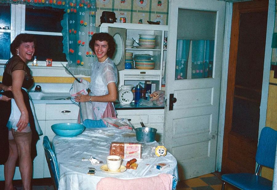 Two women smile and laugh in a colorful vintage kitchen, drying dishes by the sink. A table in the foreground holds food, dishes, and baking items, with cheerful decor and shelves of plates and cups in the background.