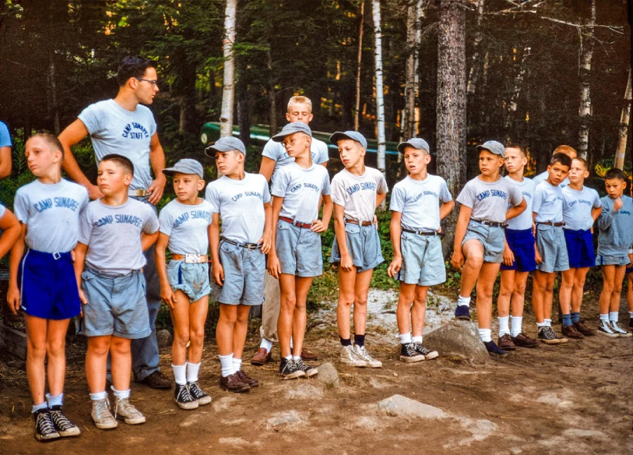 A group of young boys in matching T-shirts and shorts stand in a line outdoors with two adult leaders, surrounded by trees at what appears to be a summer camp.