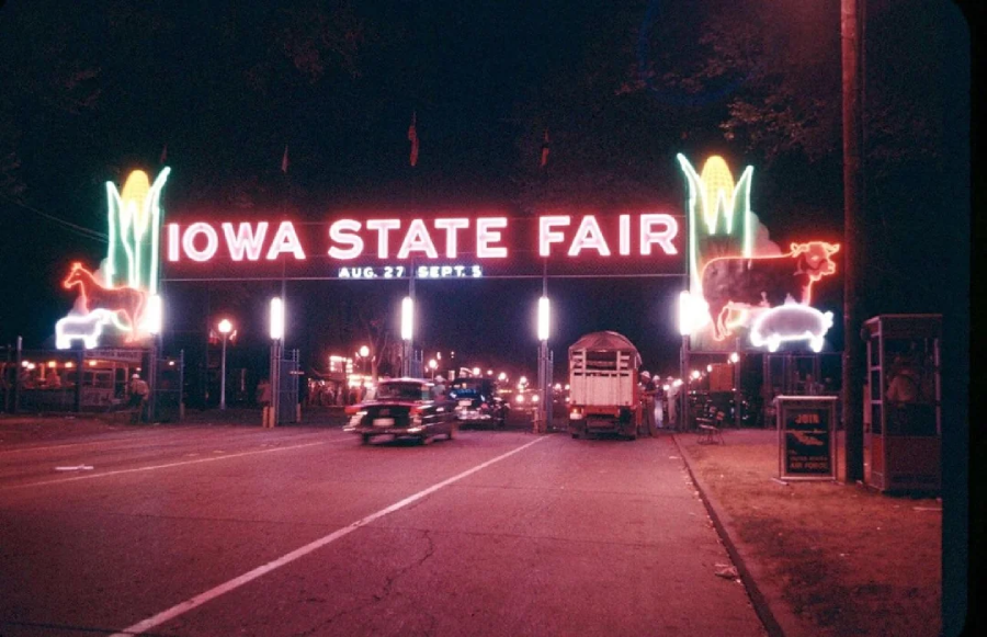 A brightly lit entrance sign for the Iowa State Fair at night, decorated with neon corn, a cow, a pig, and a horse. Cars and trucks are driving under the sign as people walk nearby.