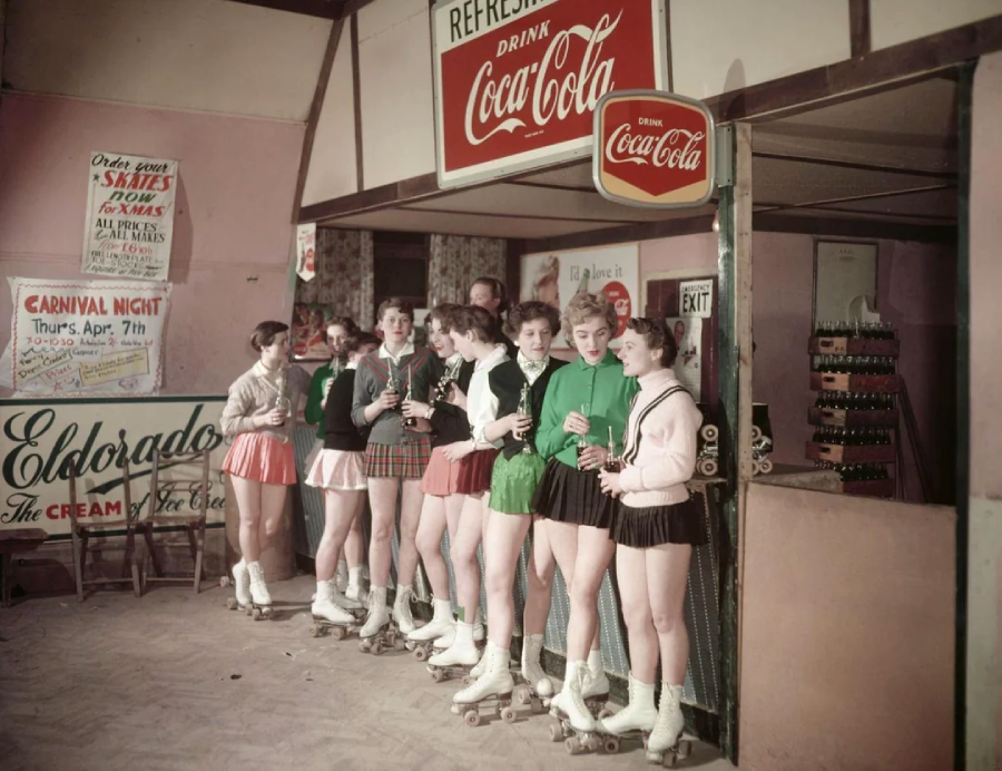 A group of young women in roller skates and colorful skirts stand in line, holding Coca-Cola bottles at a refreshment stand in a skating rink, with vintage signs in the background.