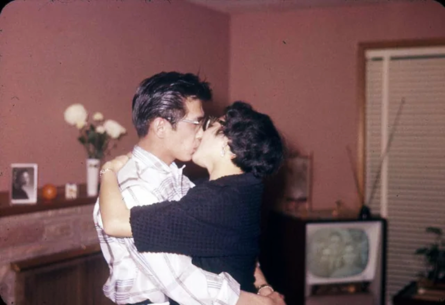 A couple embraces and kisses in a warmly lit living room with pink walls, a fireplace with flowers and a framed photo, and an old-fashioned television with a blurred image in the background.