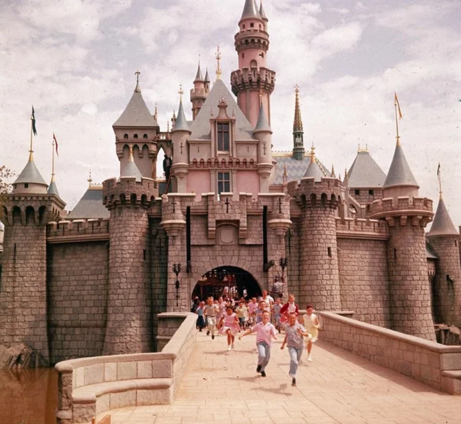 A group of excited children and adults run across the stone bridge toward the entrance of Disneyland’s Sleeping Beauty Castle on a bright, sunny day. The castle’s towers and flags are visible in the background.